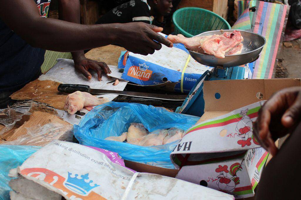 Un vendeur pointe un morceau de poulet congelé dans une poêle sur un marché informel en Afrique de l’Ouest. Cages et emballages bruns en arrière-plan. Image réelle tirée de l’article Big Chicken : le poulet importé à bas prix qui détruit les filières locales.