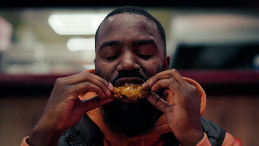 Mo Gilligan, auteur du documentaire Big Chicken sur Netflix, est en train de déguster un poulet frit gras et croustillant dans un fast-food. Yeux clos, visage expressif et concentré sur le goût. Style street-food réaliste, éclairage chaud, ambiance intime et immersive comme dans le film.