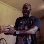 Mo Gilligan tient un poussin d’élevage industriel dans ses mains, visage illuminé par l’émotion. T-shirt noir à motif chien, bracelet doré et montre. Style réaliste, lumineux et humain comme une photo de presse du documentaire Big Chicken sur Netflix.