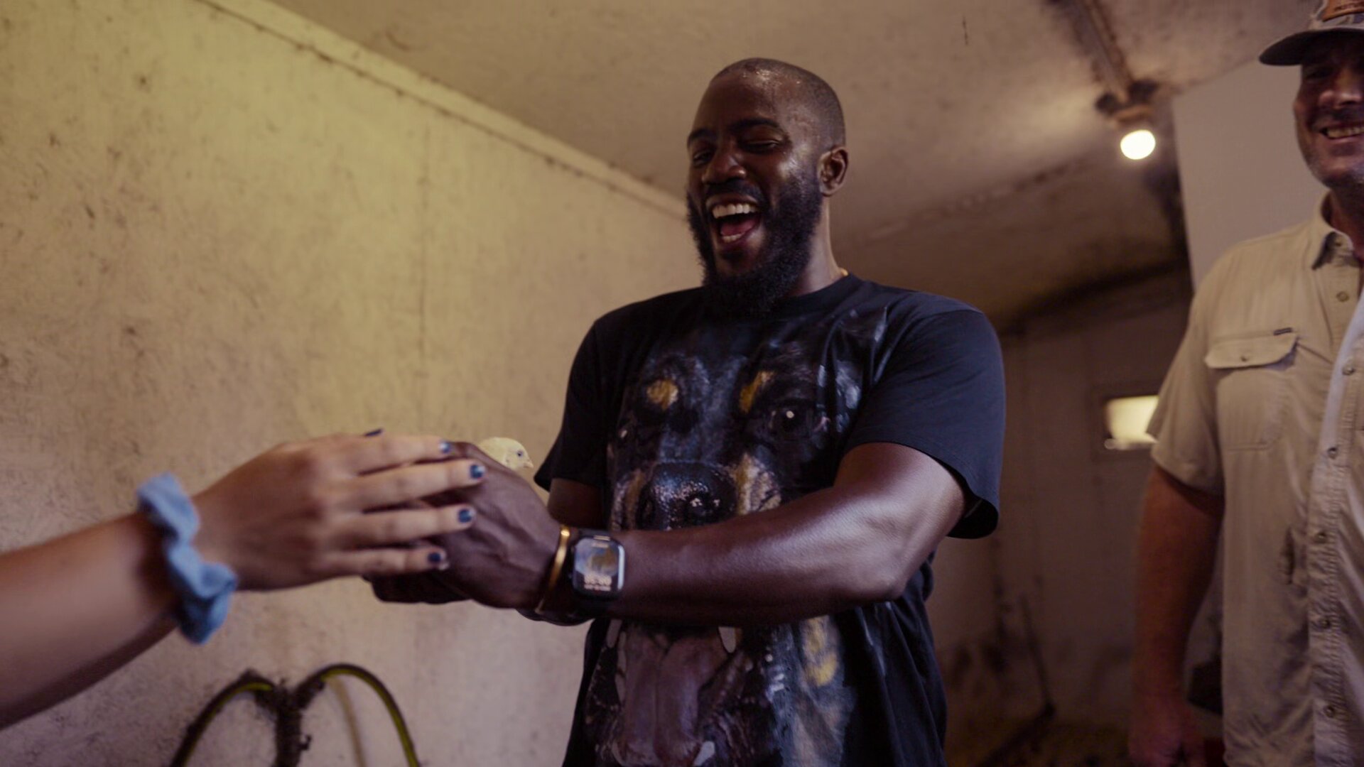 Mo Gilligan tient un poussin d’élevage industriel dans ses mains, visage illuminé par l’émotion. T-shirt noir à motif chien, bracelet doré et montre. Style réaliste, lumineux et humain comme une photo de presse du documentaire Big Chicken sur Netflix.