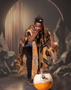 Sako Wana, chanteur burkinabé du projet Djank'Afró, vêtu d'une tenue traditionnelle aux motifs ocre et noir, jouant de la guitare calebasse devant une gourde décorée, pour le concert Sagaly 2026 à Vil