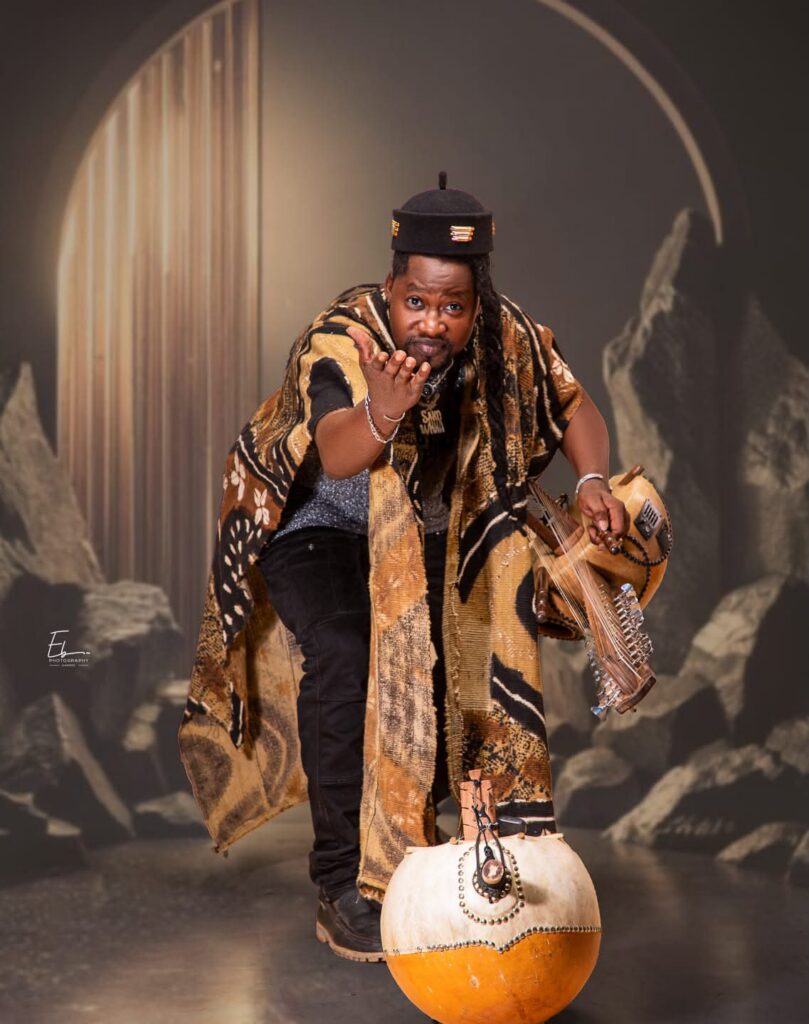 Sako Wana, chanteur burkinabé du projet Djank'Afró, vêtu d'une tenue traditionnelle aux motifs ocre et noir, jouant de la guitare calebasse devant une gourde décorée, pour le concert Sagaly 2026 à Vil